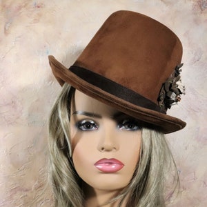 May include: A brown felt top hat with a black band and a decorative metal embellishment on the side. The hat is tilted at an angle on a person's head.