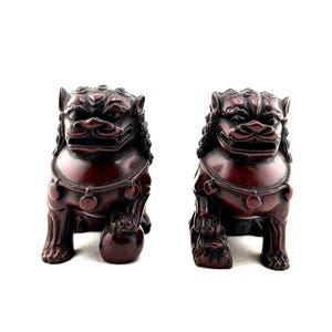 May include: Two matching dark red Foo Dog statues. Each statue has a detailed mane, a collar with circular accents, and a small ball under one paw. The statues are set against a white background.