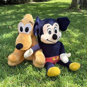 May include: Two plush Disney characters, Pluto and Mickey Mouse, sit on green grass. Pluto is yellow with black ears and a black nose. Mickey Mouse is wearing a navy blue outfit with yellow shoes and red shorts. The plush toys are in a grassy outdoor setting.