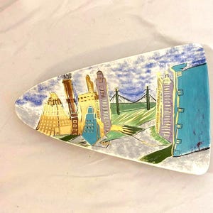 May include: A ceramic serving platter with a cityscape design. The artwork features colorful buildings, a bridge, and a cloudy sky. The platter is shaped like a long, rounded triangle.