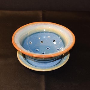May include: A blue and brown ceramic colander with a matching saucer. The colander has a ring of holes around the top and a light blue glaze.