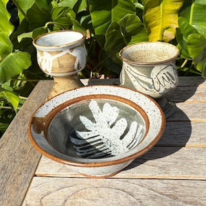 May include: Three handmade ceramic pieces: a bowl with a leaf design, and two small pots. The bowl has a brown rim and a gray interior with a white leaf design. The pots are beige and brown with abstract designs.