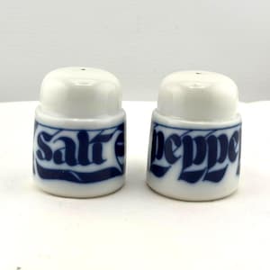 May include: A pair of white ceramic salt and pepper shakers. Each shaker has a rounded top and a cylindrical base. The words "salt" and "pepper" are written in a dark blue, stylized font around the base of each shaker.