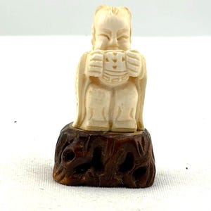 May include: Ivory-colored figurine of a seated figure with detailed clothing and facial features. The figurine is mounted on a dark brown, intricately carved wooden base. The figure appears to be holding something in its hands.