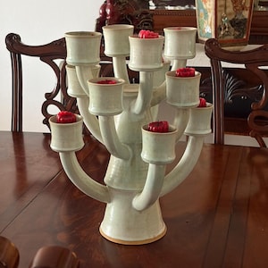 May include: A light green ceramic candelabra with multiple arms, each holding a red candle. The candelabra is placed on a dark wooden table. The design is reminiscent of a tree, with the candle holders as branches.