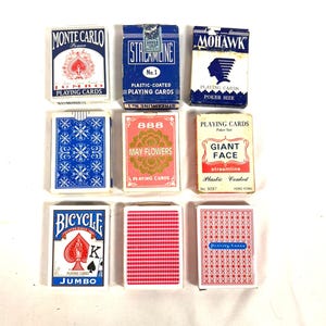 May include: Nine vintage playing card decks are displayed, each with unique designs and text. The decks feature various colors, including blue, red, and white. Some decks have text such as "Monte Carlo," "Bicycle," and "May Flowers."