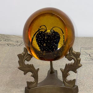 May include: A decorative amber glass paperweight with a black and gold design, resting on a brass stand. The paperweight has a spherical shape and features a dark, textured interior design. The brass stand has an ornate, vintage design.