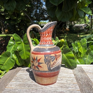 May include: A ceramic pitcher featuring a tan base with hand-painted designs in red, black, and white. The neck displays a repeating spiral pattern, while the body showcases a sun motif, a tree, and a bird, all framed by horizontal bands.