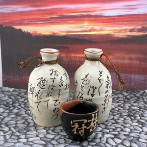 May include: Two off-white ceramic sake bottles with brown twine tied around the necks, featuring black Japanese calligraphy. A small, round, black ceramic cup with a reddish-brown interior and gold-colored Japanese characters sits in front.