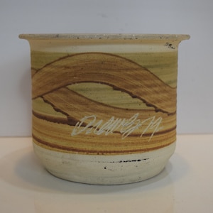 May include: A cylindrical ceramic pot with a cream-colored base and a light brown rim. The body features a wavy, abstract design in shades of brown and tan. The artist's signature is visible in white.