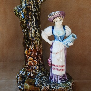 May include: A ceramic vase shaped like a tree trunk with a woman figurine holding a pitcher. The vase is decorated with brown, green, and yellow glaze. The woman is wearing a white dress with a purple skirt and a pink hat.