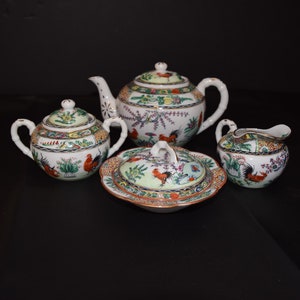 May include: A white porcelain tea set with a rooster design. The set includes a teapot, creamer, sugar bowl, and lidded dish. The design features roosters, flowers, and leaves in shades of red, green, and yellow.