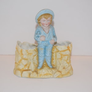 May include: A porcelain figurine of a boy wearing a blue hat and overalls standing on a yellow and white ceramic planter.