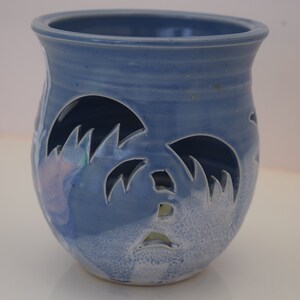 May include: A blue ceramic candle holder with a cutout design of palm trees. The holder has a white glaze and a light blue glaze.
