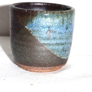 May include: A cylindrical ceramic cup featuring a two-tone glaze with a dark brown base and a textured, mottled blue upper section. The bottom rim is unglazed, revealing a coarse, reddish-brown clay texture, while the interior is dark brown.