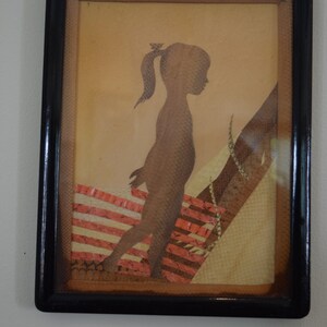 May include: A framed silhouette of a girl with a ponytail, made from textured paper in shades of brown, beige, and pink. The silhouette is set against a background of layered paper with a striped pattern.