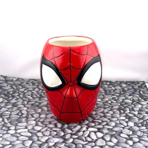 May include: A red ceramic mug shaped like Spider-Man's mask, featuring white eye shapes and black outlines. The mug has a glossy finish and is set against a neutral background.