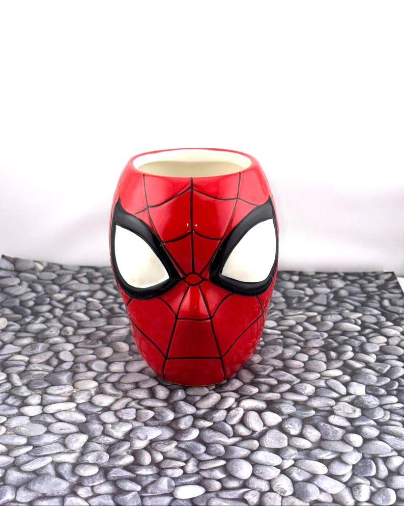May include: A red ceramic mug shaped like Spider-Man's mask, featuring white eye shapes and black outlines. The mug has a glossy finish and is set against a neutral background.