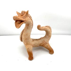May include: A terracotta-colored ceramic dragon figurine. The dragon has a long neck, a curved tail, and detailed features, including a textured mane and decorative etchings. The dragon is standing on four legs and has an open mouth.