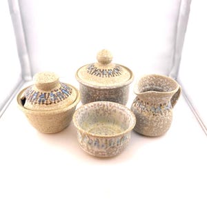 May include: A ceramic serving set with a speckled beige finish and blue and brown geometric accents. The set includes a lidded sugar bowl, a small creamer pitcher, a small open bowl, and a lidded bowl with a spoon slot.