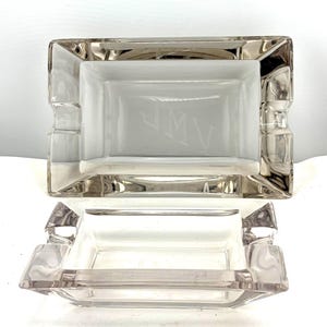 May include: Two rectangular glass ashtrays. The top ashtray has a silver rim and the initials "JMV" etched into the center. The bottom ashtray is a clear glass with a rectangular shape and a small lip on each side.