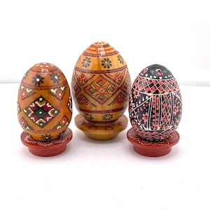 May include: Three decorative wooden Easter eggs with intricate painted designs. The eggs are in shades of brown, yellow, and black, each with unique patterns and colors. They are displayed on small, round stands, adding to their ornamental appeal.