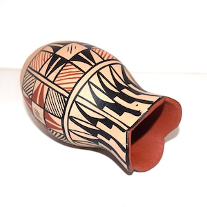 May include: A handmade, beige-colored pottery vase with a flared opening. The vase features geometric patterns in black, red, and brown. The interior of the vase is a reddish-brown color. The vase is likely used for decorative purposes.
