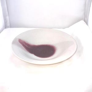 May include: A small, white ceramic dish with a dark purple, abstract design. The design is a curved shape that covers a portion of the dish's interior. The dish is round and has a slightly raised edge.