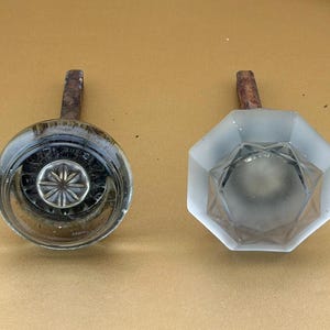 May include: Two door knobs with metal spindles. The left knob is circular, clear glass with a central starburst pattern. The right knob is octagonal, frosted glass with a faceted geometric design. Both knobs feature metallic hardware components.