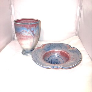 May include: A ceramic goblet and a small dish with a matching color scheme. The goblet is a tall, footed vessel with a flared rim, featuring a blend of blue, red, and white hues. The dish has a similar color palette and a decorative, scalloped edge.