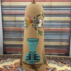 May include: A tan, bell-shaped ceramic ornament featuring a sand-textured surface. The design includes a painted figure with a white mask and green accents, positioned above a turquoise and black patterned vase, with small desert plants at the base.