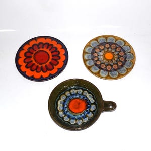 May include: Three ceramic pieces with floral designs. One is a round coaster with an orange and red flower pattern. Another is a round coaster with a blue, brown, and orange flower pattern. The third is a small dish with a handle.