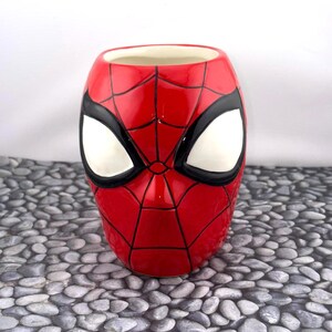 May include: A ceramic mug shaped like Spider-Man's mask. The mug is primarily red with black webbing and black outlines around the white eye shapes. The interior is a cream color.