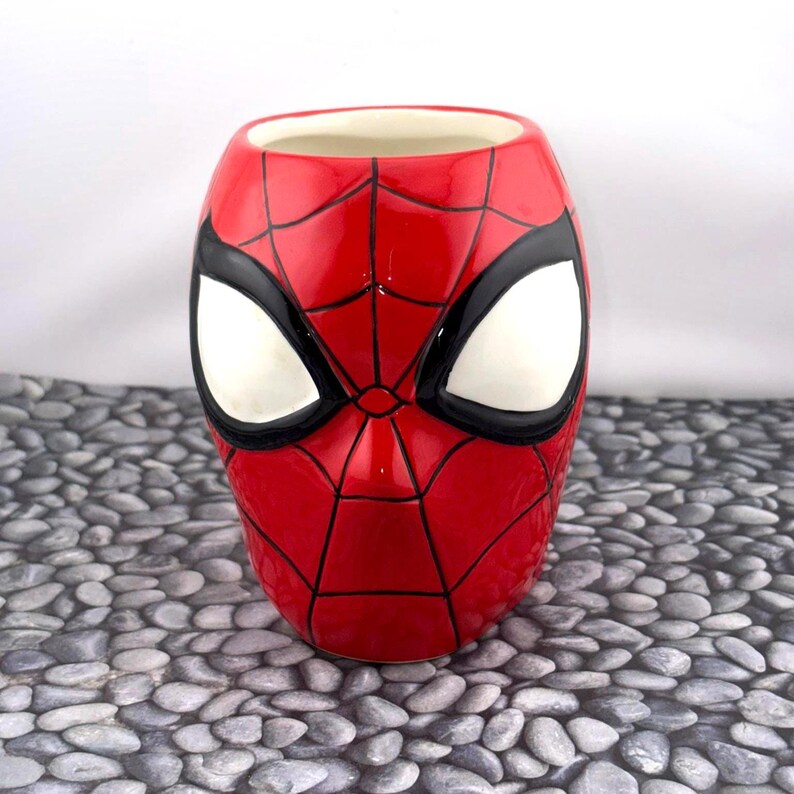 May include: A ceramic mug shaped like Spider-Man's mask. The mug is primarily red with black webbing and black outlines around the white eye shapes. The interior is a cream color.