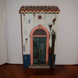 May include: A miniature model of a building with a red tile roof, a pink door with a wrought iron window, and a white facade with blue and brown accents. The building has a number "46" on the side.
