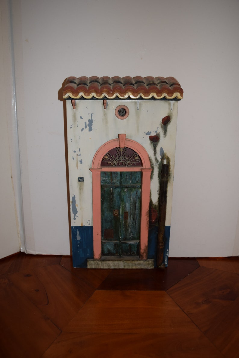 May include: A miniature model of a building with a red tile roof, a pink door with a wrought iron window, and a white facade with blue and brown accents. The building has a number "46" on the side.