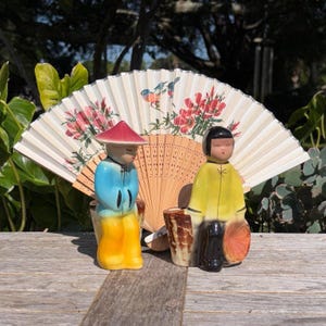 May include: A decorative display featuring two ceramic figurines dressed in traditional attire, with a folding fan in the background. The fan is cream-colored with floral and bird designs. The figurines are in blue, yellow, and black.