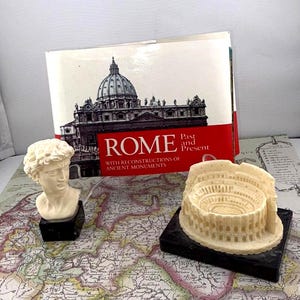 Op de afbeelding: Een stilleven met een boek getiteld "ROME" met een afbeelding van een gebouw op de omslag. Twee kleine sculpturen, een buste en het Colosseum, staan op een kaart. De sculpturen staan op zwarte sokkels.