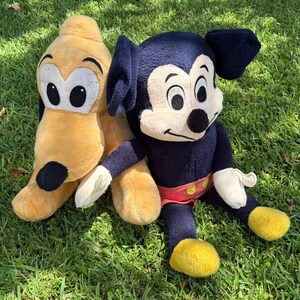 May include: Two plush Disney characters, Mickey Mouse and Pluto, sit on green grass. Mickey Mouse is dark blue with a cream face, black ears, and yellow shoes. Pluto is yellow with black ears and a black spot.