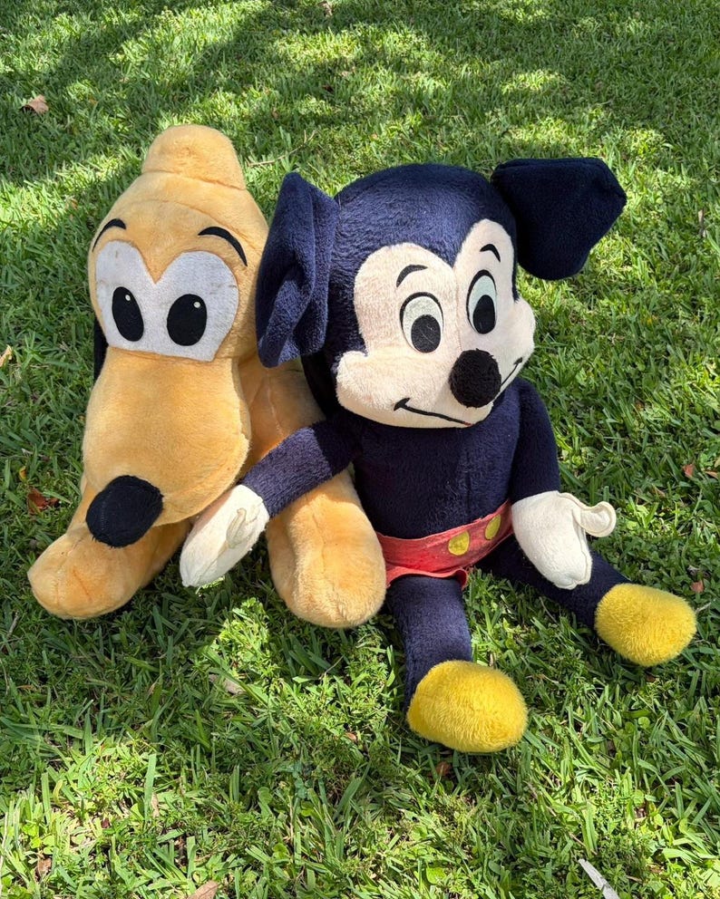 May include: Two plush Disney characters, Mickey Mouse and Pluto, sit on green grass. Mickey Mouse is dark blue with a cream face, black ears, and yellow shoes. Pluto is yellow with black ears and a black spot.