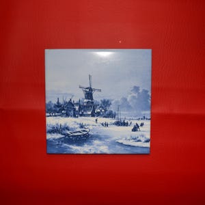 May include: A square, blue and white ceramic tile featuring a winter landscape scene. The image depicts a windmill, houses, a frozen body of water, and figures ice skating. The tile has a glossy finish and is set against a red background.