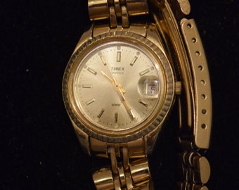 May include: A gold-toned Timex quartz wristwatch with a gold-tone metal band. The watch face is white with black markings and a date window.