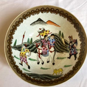 May include: A decorative ceramic plate with a detailed hand-painted scene. The plate features figures on horseback, mountains, and a border with a repeating pattern. The color palette includes brown, white, green, yellow, and red.