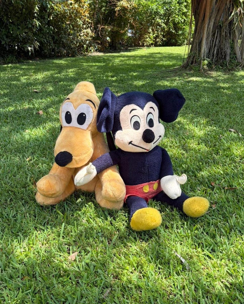 May include: Two plush Disney characters, Pluto and Mickey Mouse, sit on green grass. Pluto is yellow with black ears and a black nose. Mickey Mouse is wearing a navy blue outfit with yellow shoes and red shorts. The plush toys are in a grassy outdoor setting.