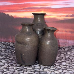 May include: Three dark brown ceramic vases with smooth, rounded bodies and flared rims. The vases are arranged in a cluster, with one taller vase positioned behind two shorter vases. Each piece features a consistent matte finish and simple form.