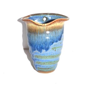 May include: A handmade ceramic wall vase with a wavy rim. The vase features a blend of blue, green, and brown hues, with a hole at the top for hanging. The exterior has a textured, layered appearance.