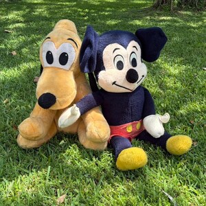 May include: Two plush Disney characters, Pluto and Mickey Mouse, sit on green grass. Pluto is yellow with a black nose and ears. Mickey Mouse is navy blue with a red shorts and yellow shoes. Both are soft toys.