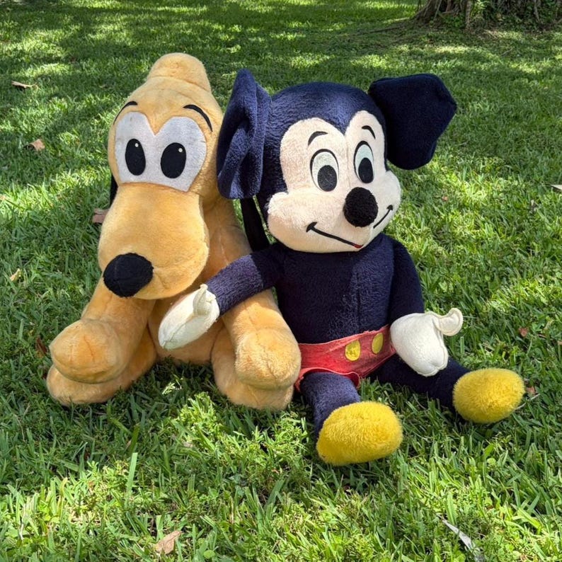May include: Two plush Disney characters, Pluto and Mickey Mouse, sit on green grass. Pluto is yellow with a black nose and ears. Mickey Mouse is navy blue with a red shorts and yellow shoes. Both are soft toys.