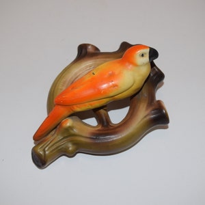 May include: A ceramic bird figurine with orange and yellow plumage perched on a brown branch. The bird has a black eye and beak. The branch is shaped like a circle.