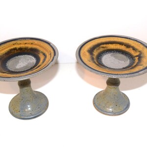 May include: Two ceramic pedestal bowls featuring a wide, shallow basin with a circular design in shades of mustard yellow, dark brown, and grey. The base of each bowl is a solid, speckled grey color with a flared, sturdy foot for stability.